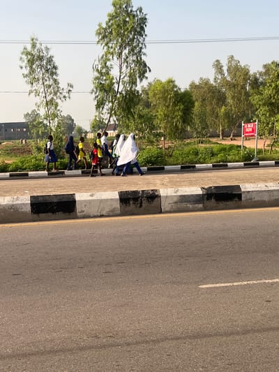 In Kporva, Nigeria, a group of school-aged children, dressed in uniforms of blue skirts, white shirts, and some with yellow or red shirts, walk along a paved sidewalk adjacent to a road. Some children wear headscarves, with one child covered in a white hijab. A red sign with white lettering is visible in the background, partially obscured by trees. The environment appears to be a semi-urban area with trees lining the background and a building visible in the distance. The sky is clear and a light blue. The road in the foreground is paved with asphalt and has visible white lines.