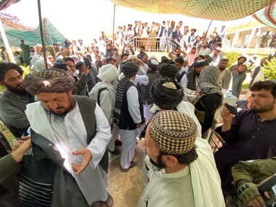 A gathering of approximately 40 people, predominantly men, takes place in a paved courtyard in Zaker-E Sharif, Afghanistan. The area is shaded by a large white tent. Most men wear traditional attire, including white shirts, loose trousers, and black or white turbans. They stand in a large group, seemingly conversing.

A man in a white shirt and white pants stands centrally, with a stoic expression. Beside him, another man with a black turban, white shirt, and black vest shares the same serious demeanor. To the central figure's left, a bearded man in a black turban and black shirt looks serious, gazing off to the side.

In the foreground, a bearded man in a black shirt and black turban, with a somber expression, operates a cell phone with his left hand. He appears to be filming or photographing the central group, with his back facing them. His right hand rests on his lap.

The blurred background reveals additional traditionally dressed men, some looking towards the camera, others away. A few women, their faces obscured but dressed in traditional clothing, are also visible in the far back under the tent. Bright sunshine indicates a warm, clear day, contributing to a calm and peaceful atmosphere.