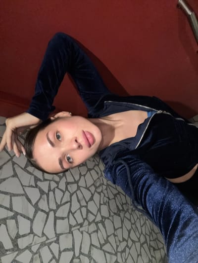 A young woman is lying on a floor with a mosaic tile pattern. She is wearing a blue velvet zip-up jacket. A red surface is visible in the background. The location may be Borok, Russia.