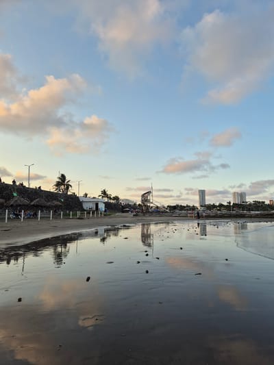 The image shows a beach in Veracruz, Mexico, at dusk. The sky is a mix of blue and clouds with pinkish hues. On the right, the wet sand reflects the sky and the city buildings in the distance. Along the beach, there is a breakwater made of rocks, palm trees, light posts, and some small buildings or stalls. In the distance, high-rise buildings mark the city's skyline. Some people are visible on the beach, but they are not the primary focus of the image. The composition is dominated by the sky and the reflections on the wet sand, creating a calm and serene atmosphere.