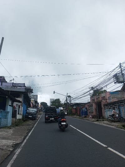 The image depicts an overcast street scene in what is identified as Ariyalai East, Sri Lanka. A two-lane asphalt road, marked with a solid white line separating directions, recedes into the distance under a uniformly grey, cloudy sky. Densely strung black utility cables traverse the upper frame, supported by multiple utility poles visible on both sides of the road.
On the left, a narrow, uneven concrete sidewalk borders a row of multi-story buildings, some appearing aged or weathered. A dark-colored small utility truck or pickup is parked partially on this sidewalk, facing away from the camera, its front end appearing low.
On the right, another row of buildings lines the street, including a prominent pinkish-brown structure featuring decorative elements on its facade. A complex network of utility wires and equipment is mounted on poles above these structures. Further down the street, a traffic light signal is visible. A person wearing a light-colored shirt is seated stationary on a classic-style motorcycle, facing left, positioned near the curb on the right side.
Vehicular traffic moves away from the camera in the center of the road. A dark-colored utility truck with Indonesian license plate "D 9345 NJ" proceeds forward. Following it is a black scooter, ridden by a person wearing a light helmet and dark jacket, also moving away from the camera. This scooter displays an Indonesian license plate "P 6914 NM".