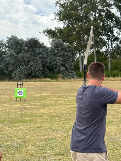 A person wearing a gray t-shirt and tan pants is shown drawing back a bow and arrow aimed at a green and blue target in a grassy field. The target is placed in the middle of the field. Trees and a cloudy sky appear in the background. The image is taken from behind the person.