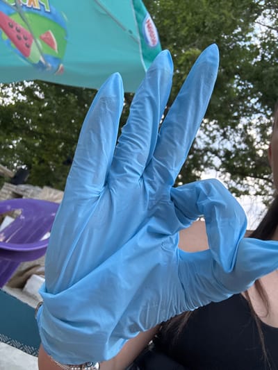 A forensic examination of the image reveals a person's right hand, clad in a light blue disposable glove, making an 'okay' gesture. The thumb and index finger are joined to form a circle, while the middle, ring, and pinky fingers are extended upwards. A metallic bracelet is visible on the wrist, partially covered by the glove. In the blurred background, an aqua-blue umbrella with a green segment, a red watermelon design, and a white 'Nestlé' logo is discernible in the upper-left quadrant. Beneath the umbrella and to the center-right, out-of-focus green foliage, characteristic of trees, is visible. A portion of a purple object, possibly a bowl or container, is located in the lower-left corner on a patterned surface. The right side of the frame shows the dark hair, ear, and a black garment of the individual holding the hand. The scene is located in Kostievo, Bulgaria.