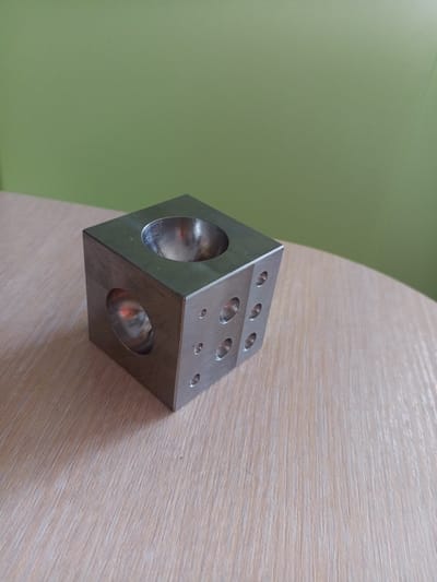 A metallic cube with multiple holes and indentations sits on a light-colored wooden table. The cube has a large circular opening on its top face and another on its left face. The front face of the cube features several smaller circular indentations. Behind the table, a plain light green wall is visible. The image is taken from a medium-angle perspective, focusing on the cube and the table surface. There are no people present in the image. The scene does not provide any specific geographical indicators to determine its location within Vilyuysk, Russia.