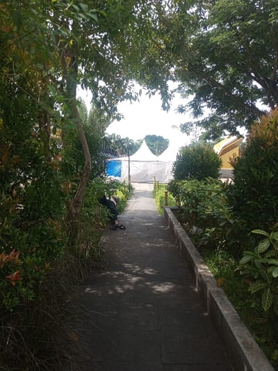 A tree-lined walkway leads toward a white tent structure in the distance. Lush greenery flanks either side of the path, creating a natural tunnel effect. A person is resting or sleeping beside the path on the left. The walkway is paved with dark gray stone, dappled with sunlight and shadows from the trees above. A yellow building can be seen to the right of the tent.