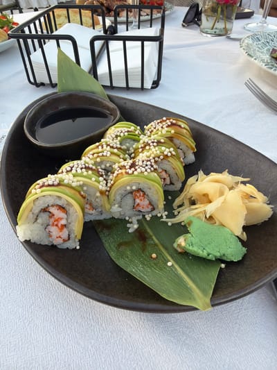 A close-up shot shows a dark oval plate with a serving of sushi rolls, pickled ginger, and wasabi on a white tablecloth. The sushi rolls are arranged in a curved line, topped with thin slices of avocado and sprinkled with small white balls. The plate also contains a small dark bowl of soy sauce and a large green leaf, possibly a banana leaf, used as a garnish. In the background, a black wire basket holds stacks of white napkins. Other elements in the background, though out of focus, suggest an outdoor dining setting, with hints of silverware and a glass. The overall scene appears to be a well-presented meal, likely at a restaurant, but the specific location in Vatra, Moldova, cannot be determined from the visual information provided.