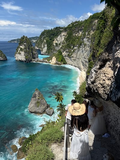 The image captures a bird's-eye view of Kelingking Beach in Pejukutan, Indonesia. The scene features turquoise waters, a white sandy beach, and towering cliffs covered in lush greenery. At the edge of the cliff, several people wearing white dresses and hats are positioned near a rope fence. The composition emphasizes the contrast between the natural landscape and the human figures.