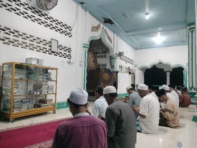 This image displays the interior of a mosque in Tumpeun, Indonesia, during a religious gathering. Approximately fourteen adult males are present, seated on the floor in rows, facing an ornate, dark wooden mihrab or minbar structure at the front of the room. Most individuals wear white or black skullcaps (kufi or peci) and various forms of traditional attire. Their posture indicates a state of prayer or contemplation.
To the left of the frame, a yellow-framed glass display cabinet with caster wheels holds numerous books stacked on multiple shelves. A white rectangular box rests on top of this cabinet. The walls are white, featuring horizontal bands of repeating dark grey or black stenciled patterns and green and white arched decorative trim that also adorns visible pillars and architectural niches. A square framed object is mounted on the upper left wall. A wall-mounted oscillating fan is visible high on the left. The ceiling is paneled in light blue and contains multiple flush-mounted fluorescent light fixtures. The floor consists of white tiles, with a segment of red patterned carpet in the immediate foreground where some individuals are seated.