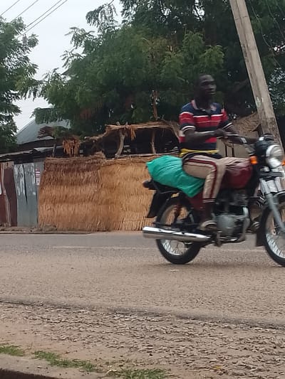 In Biliri, Nigeria, a man rides a motorcycle on a paved road. He wears a red and blue striped shirt and tan pants. A green bag is behind him on the motorcycle. In the background are trees, a utility pole, and a structure with corrugated metal and straw walls. The sky appears overcast.