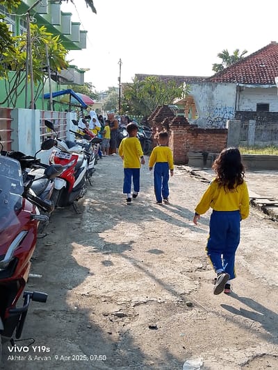 The image shows a street in Kecamatan Gegesik, Indonesia. A group of children are walking down the street, in the direction of the camera. One girl is wearing a yellow shirt and blue pants and walking with a slight backward lean. The other two children are both wearing yellow shirts and blue pants and walking in the same direction. The children are walking between a line of parked motorcycles on the left and brick walls on the right. The ground is cracked and uneven, and the street is shadowed. There are several adults and children standing in the background, and they appear to be interacting with each other. There is a house on the right side of the street, and the walls are covered in graffiti.