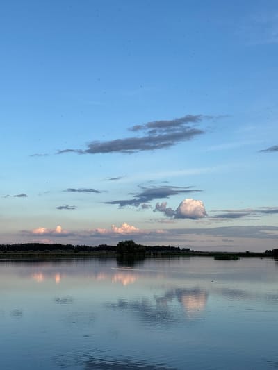 The image depicts a body of water, likely a lake or wide river, under a partly cloudy sky. The water surface reflects the sky and clouds, creating a mirrored effect. A line of trees and vegetation is visible on the far bank. There are also many specks in the sky, possibly bugs. The sky transitions from a clear blue to a lighter, duskier hue near the horizon. The scene appears peaceful and serene. While the location is specified as Khryashchevka, Russia, this cannot be verified from the image itself, as no identifiable landmarks or features are present. The image appears to be a landscape shot.