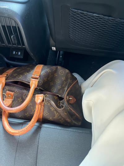 A Louis Vuitton duffel bag is nestled between the back seat and the center console of a car. The bag is brown with the iconic LV monogram pattern and has tan leather handles. A pair of white pants or leggings are visible above the bag. The car's interior includes a mesh pocket, a USB charging port, and a textured gray seat.