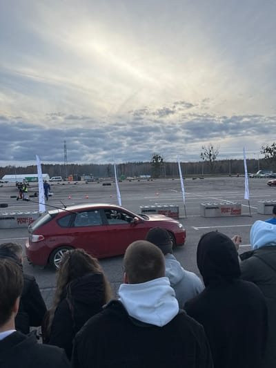 The image displays an outdoor automotive event taking place in Wolica, Poland, on a large, dark gray asphalt surface. The sky overhead is overcast with multiple cloud layers. A red, five-door Subaru Impreza hatchback is prominently positioned horizontally in the mid-ground on the asphalt; a driver is visible within the vehicle. In the foreground, a group of approximately 6-8 individuals, observed from behind, stand facing the event area, dressed in casual, dark attire. The event space is marked by multiple white vertical flags and gray concrete barriers, both branded with "MANNOL" and "FUNDACJA FUN-DRIVE." Orange traffic cones and scattered tires are visible on the asphalt, outlining a potential course. In the background, a dense line of bare deciduous trees forms a horizon, with a communication tower visible above the tree line to the left. Further in the background, a white tent and several white vans are present, along with individuals, some wearing high-visibility vests, near a scooter-like device.