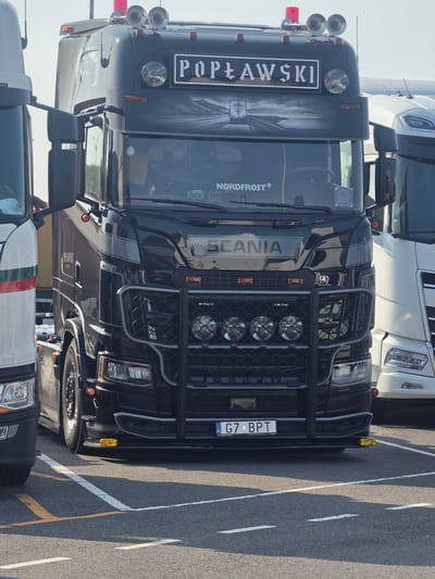 A black Scania semi-truck is parked in an outdoor lot, possibly a truck stop or industrial area, in Maasvlakte Rotterdam, Netherlands. The truck features a custom paint job with "POPLAWSKI" emblazoned on the front above the windshield. It also has a bull bar with four auxiliary lights and dual horns mounted on the roof, accompanied by a red flashing beacon. The truck's license plate is visible and reads "G7 BPT". To the left and right of the Scania are other trucks, one predominantly white and another partially visible white truck with green and red stripes on its side. The ground is asphalt with painted parking lines. The sky is clear and bright.