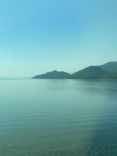 The image depicts a serene lake scene with mountains in the background. The sky is a clear, light blue, fading to a slightly lighter hue near the horizon. The lake itself is a tranquil expanse of calm, greenish-blue water, reflecting the subtle colors of the sky. Gentle ripples disturb the surface of the water, particularly in the middle ground, suggesting a slight breeze. The foreground of the lake reveals the submerged bottom, covered with patches of green algae or aquatic vegetation.
In the distance, a series of rolling, green-hued mountains form the horizon. The mountains are covered in lush vegetation and their shapes are rounded, with softer slopes rather than sharp peaks. The leftmost mountains are fainter, appearing hazy due to atmospheric perspective. A small, dark object, possibly a bird or a floating piece of debris, is visible on the water's surface in the middle right portion of the image.
The scene is devoid of any human presence or man-made structures. The overall impression is one of natural beauty and peacefulness. While the prompt mentions the city of Kruševica, Montenegro, this specific image does not contain any identifiable landmarks or clues that definitively place it in that particular location. However, the natural landscape of mountains and a large body of water is consistent with many areas in Montenegro.