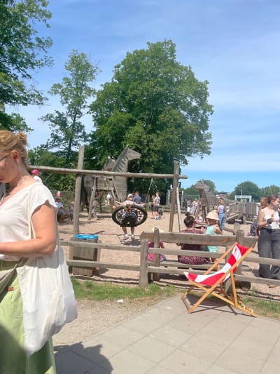 This outdoor photograph, taken in Wysoka, Poland, depicts a lively playground or recreational park under a bright, sunny sky. The image is oriented vertically. In the foreground, partially visible on the left, is a person with blonde hair and glasses, wearing a white sleeveless top.
The central and middle ground of the scene features an extensive wooden playground structure, possibly designed to resemble a fort or village. Children are actively engaged with the equipment, including a large, circular nest swing where one child is seated. Several adults are also present, standing, sitting on wooden benches, and supervising the children.
The ground comprises dirt, gravel, and paved concrete pathways. A wooden fence or barrier runs along the path, separating it from the playground area. To the right, a wooden deck chair with a distinct red and white striped fabric is visible, positioned beside the pathway.
Tall, lush green trees frame the park, their leaves vibrant under the clear blue sky, which shows only a few very faint, wispy clouds. The strong sunlight casts prominent shadows, indicating it is midday or early afternoon. The overall atmosphere is one of leisure and enjoyment in an outdoor setting. No discernible text is visible on any signs or objects.