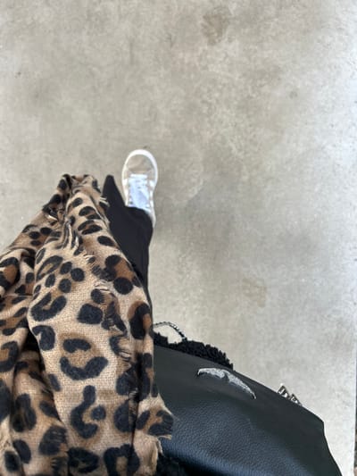The image displays an overhead, close-up perspective of a partially visible individual standing on a light grey, textured ground surface in Saint-Étienne, France. The lower right leg of the individual is dressed in black trousers, with the right foot encased in a white sneaker featuring white laces. A tan and black leopard print scarf, exhibiting frayed edges, is draped across the lower left quadrant of the frame. Adjacent to the scarf and below the visible leg, a black textured handbag is present, adorned with a distinct silver-toned winged emblem on its exterior. No other subjects or specific environmental identifiers are visible within the frame.