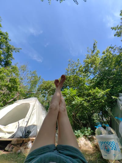 In the city of Utios, Ukraine, a person is lying down outdoors, with their legs crossed and raised towards a bright blue sky dotted with a few wispy clouds. The person is wearing dark green shorts. In the background, a white tent is set up, resting on a wooden platform. The tent is open, revealing its interior. Surrounding the tent and the person are lush green trees and foliage, suggesting a natural, possibly wooded environment. To the right of the tent, there are several plastic bottles, two of which are large and clear with blue caps, and a white bucket with blue checkered pattern and a drawing of a girl. The ground appears to be covered in grass and some scattered rocks and debris. The overall scene depicts a moment of relaxation in an outdoor setting.