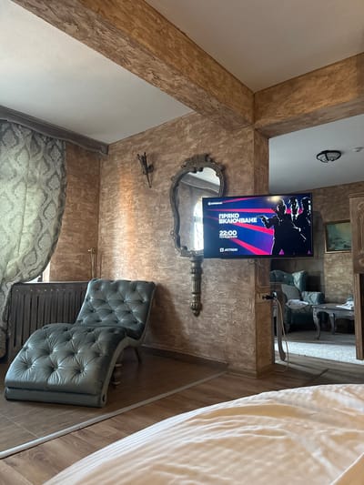 The image depicts the interior of a hotel room in Ognyanovo, Bulgaria. A flat-screen television is mounted on the textured, beige wall, displaying a program with Bulgarian text and silhouettes of three individuals holding firearms. Below the television, a dark grey chaise lounge with button tufting is positioned on a wooden floor. A patterned grey curtain hangs on the left side of the room, partially obscuring a radiator. A decorative, ornate mirror is mounted on the wall above the chaise lounge, and a wall sconce is visible to its left. In the background, through an open doorway, a glimpse of another room is visible, containing a sofa, a table, and a framed painting on the wall. The lighting suggests an indoor environment, possibly during the day, with natural light filtering through the curtain.