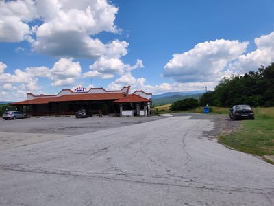 This is a wide-angle, eye-level shot of a roadside establishment on a sunny day in Osikovitsa, Bulgaria. The building, with a red-tiled roof and white facade, appears to be a restaurant or cafe, with outdoor seating and a small kiosk. The name "БИСТРО ПЕРЛА" (BISTRO PERLA) is visible in blue lettering on the building's facade.
The scene is set against a backdrop of rolling green hills and mountains under a bright blue sky dotted with fluffy white clouds. The foreground consists of a cracked asphalt parking lot and road, suggesting a well-trafficked area. Several cars are parked in front of the establishment, including a silver SUV and a dark-colored sedan. A blue trash bin is also visible.
The lighting suggests it is daytime, likely mid-morning or afternoon, with ample sunlight casting shadows. The overall atmosphere is one of a typical rural stopover, offering a place to rest and refuel amidst scenic Bulgarian landscape. There are no people prominently featured, though a few figures are vaguely discernible near the building entrance, their activities unclear. The weather appears pleasant, suitable for outdoor activities.