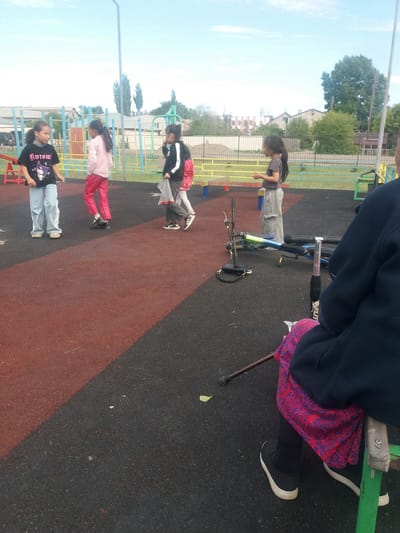 The image shows a group of children in an outdoor playground in what appears to be Бородулиха ауылдық округі, Kazakhstan. The playground has a red and black rubber surface. There are four children standing in a line, appearing to be engaged in an activity or game. To the upper right, a person is partially visible, sitting on a piece of playground equipment, possibly a seesaw, with a bicycle nearby. A cane or walking stick is visible near the person seated on the equipment. The environment includes a fence, buildings, trees, and light poles, indicating a residential area.