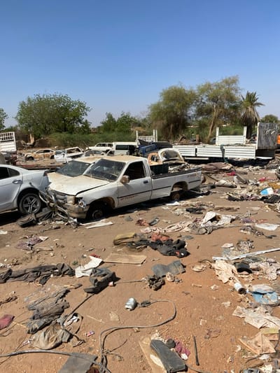 The image shows a scene of destruction in Tayba Alhasanab, Sudan (latitude: 15.36, longitude: 32.528). There are several vehicles, including a white pickup truck in the foreground, and several other vehicles in the background. The vehicles appear to be damaged. The ground is covered in debris and rubble. There are trees and bushes in the background. The image suggests that there has been a recent conflict or disaster.