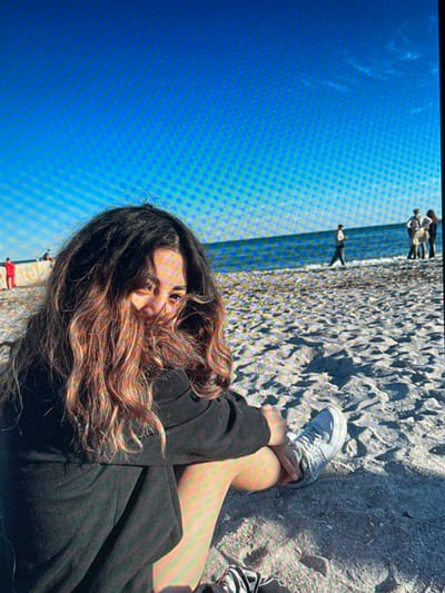 The image shows a young woman sitting on a sandy beach, with her hair covering much of her face. She is wearing a black hoodie and light-colored sneakers. The background features a bright blue sky, the ocean, and other people in the distance. The lighting suggests it is daytime, with shadows cast on the sand. The location of Chotěšov, Czechia, is not supported by any visual evidence in the image.