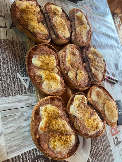 A table-level, eye-level, close-up, medium shot captures a spread of toasted bread slices topped with melted cheese and sprinkled with spices, arranged on a wooden board placed on a table with a patterned surface. The toast slices are cut lengthwise from a baguette-style loaf and have a dark brown crust. The cheese is a pale yellow and appears melted and slightly browned in places, with visible specks of seasoning. Some slices show a reddish-brown spread beneath the cheese, possibly a meat paste or pâté. The background features a textured, patterned tablecloth or placemat in shades of brown, beige, and white. A portion of a white plate is visible at the bottom left, and a black electronic device with a silver button is partially visible on the right. The lighting is natural and appears to be coming from above and to the left, casting soft shadows. The scene is indoors, suggesting a meal preparation or serving setting, and there are no visible people. The location is the village of Karabunar, Bulgaria.