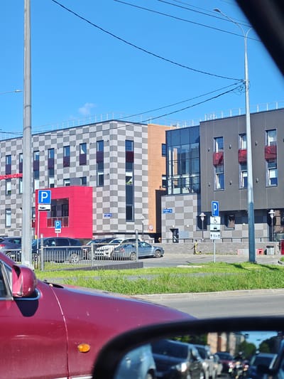 The image shows a modern building in Petrozavodsk, Russia, with a checkered facade of grey and white squares. The building has large windows and balconies with red railings. In front of the building, there is a red-accented entrance area with parking signs. Several cars are parked in the foreground, and a yellow electric scooter is visible on the sidewalk. The sky is clear blue. The scene appears to be a commercial or office building located in an urban area.