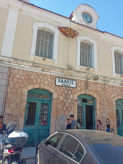 The image captures the exterior of a train station in Chalkis, Municipal Unit of Anthidon, Greece. The station building features a cream-colored upper facade with arched windows and white trim, topped by a red-tiled roof and a decorative clock. Below the windows, the building is constructed with a distinctive hexagonal stone pattern. A sign prominently displays "ΧΑΛΚΙΣ" and "HALKIS" in black lettering on a white background. Two teal-colored doors with ornate metal grilles are visible, leading into the station.
In front of the station, several people are present. Two young men and a woman with headphones are standing near an open doorway. Another woman, wearing a black tank top and jeans, is talking on her phone. To the left, a motorcycle with a white top box is parked on the sidewalk. A dark-colored car is partially visible in the foreground, obscuring some of the lower portion of the station. Graffiti is present on the stone facade of the building. The sky is clear and blue, indicating a sunny day.