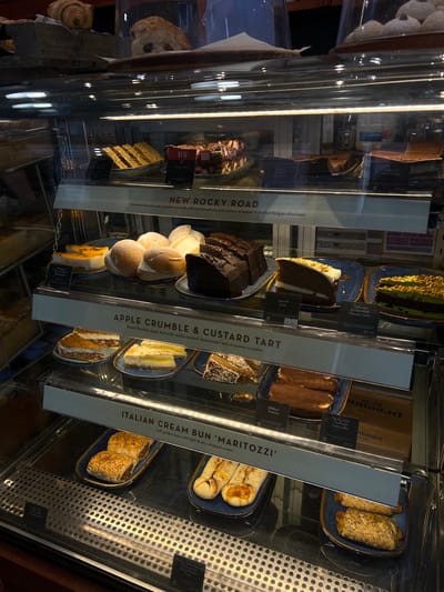 The image captures a refrigerated display case in a food establishment in Belfast, United Kingdom. The case contains a variety of desserts and baked goods arranged on multiple shelves. Signs are placed in front of each item, identifying them and their prices.
The desserts include "New Rocky Road," "Apple Crumble & Custard Tart," "Italian Cream Bun 'Maritozzi'," "Sticky Toffee Pudding Cheesecake," "Pistachio & Chocolate Mousse Cake," "Carrot Cake," "Italian Tiramisu," "Oryvh Pork Sausage Roll Folded," "Caramol Shortbread," and "Rocky Road Chocolate Brownie."
The environment appears to be well-lit and clean. The display case is made of glass, offering a clear view of the desserts inside. The metallic frame of the case reflects light, adding to the brightness of the scene. A cooling vent is visible on the right side.