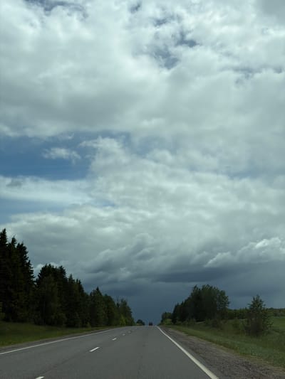 A road runs from bottom right to top center, with a double yellow line marking its center. Dense foliage lines the right side of the road, transitioning into a tree line further away. A vehicle with red lights is visible in the distance on the road. The sky is mostly cloudy, with a small patch of blue visible. A heavy, dark cloud bank stretches down from the top edge of the frame on the left. The photo was likely taken on a road in the Юзуфоўскі сельскі Савет.