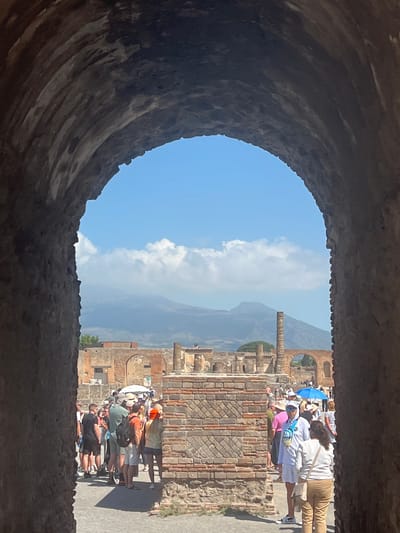 This is a photograph of the ancient city of Pompeii, Italy, taken from within a stone archway. The archway frames a view of the ruins, with a large gathering of tourists visible in the middle ground. Beyond the ruins, a hazy mountain range, likely Mount Vesuvius, can be seen under a bright blue sky dotted with white clouds. The tourists, dressed in casual summer attire, appear to be exploring the archaeological site. Some are holding umbrellas for shade, suggesting a warm, sunny day. The ruins consist of brick walls and columns, characteristic of Roman architecture. The foreground of the image is dominated by the dark, textured stone of the archway, creating a sense of depth and perspective.