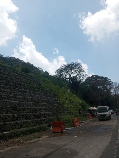 The image shows a section of roadway adjacent to a terraced hillside in an unspecified location, likely not Nawng Hkar, Myanmar. The hillside has been reinforced with wire mesh containing rocks. A white truck is parked on the side of the road. Orange traffic barriers are present, with debris scattered along the roadside. A person is sitting near a small stall under an umbrella. A motorcycle is visible in the background. The sky is blue with scattered clouds.