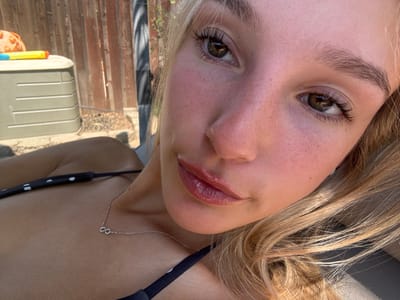 The image displays a close-up, first-person perspective, consistent with a selfie, of a female subject in an outdoor residential setting. The subject, a young adult with blonde hair and light-colored eyes (hazel), occupies the majority of the frame, looking directly forward. Her facial skin exhibits a reddish flush, consistent with sun exposure, and her lips appear glossy and slightly parted. She is wearing a black swimsuit top with white polka dots. A thin, silver-colored necklace with an infinity symbol pendant rests on her sternum.
The background is out of focus, revealing elements of a residential backyard. To the subject's left, a light-grey, rectangular plastic storage container is visible. On its top surface, a yellow and blue toy, consistent with a water pistol, and a partially visible orange, spherical object are present. Behind these objects and the subject, a weathered brown wooden privacy fence is discernible. The ground immediately adjacent to the storage container appears to be dry soil or gravel. Strong, natural light indicates a daytime setting. The scene is situated within a residential backyard in Rocklin, United States.