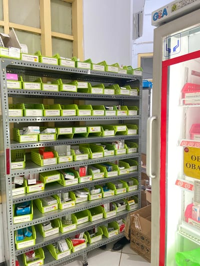 The image shows an interior view of a pharmacy, likely in Margoyoso, Indonesia, showcasing rows of medication organized in green plastic bins on metal shelving units. The scene is brightly lit, suggesting it's daytime. The focus is on the organized storage of pharmaceuticals, with various boxes and packets clearly visible.
Prominent text includes brand names and dosages such as "GEA", "ROCULAX INJ", "OXYT", "OB", "OBA", "QUEENVIT TAB", "NITROKAF RETARD FORTE", "OBESLIM 10 MG KAPSUL", "PROSTACOM", "RANTIN 150 MG TAB", "ENAX", "RETIVIT PLUS", "SYALOX 300 P TAB", "TIRIZ", "TOFEDEX 25 MG TAB", "TRICHODAZOL 500 MG TAB", "UROGETIX 100 MG", "UPERIO 50 MG L", "V-BLOC 6,25MO", "RIFAMPICIN DO M", "PIRACETAM 800 MG TAB", and "VILDAGLIPTIN SO ma". There are also "HIGH ALERT" labels visible on some products. A sign with the text "CFC DAN HOFC" and another indicating "RUMAH Apotik R HRSYAP 2/8" provide further location context. The overall impression is one of a well-stocked and systematically managed pharmacy.