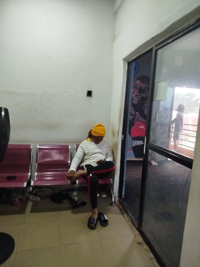 The image shows a person sitting on a row of maroon chairs in what appears to be a waiting area or lounge. The individual is wearing a yellow beanie, a white patterned sweater, and black pants with red stripes. They are looking down at a mobile phone. Scattered on the floor in front of the chairs are several pairs of shoes and sandals. To the right of the waiting area is a large glass sliding door, revealing a glimpse of the outside. Through the glass, another person can be seen standing and looking outwards, with a railing and some structures visible in the background. There is a mural of a boxer's face on the wall next to the door. The lighting suggests it could be daytime. The overall scene conveys a sense of waiting or resting.
