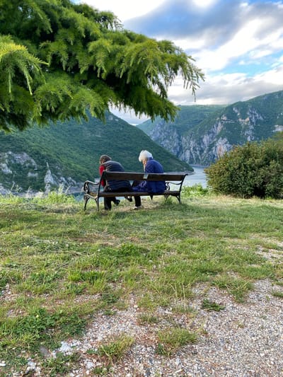 The image captures a picturesque landscape view from an elevated vantage point in Plužine, Montenegro. Two people are seen sitting on a dark wooden and metal park bench, with their backs to the camera, gazing out at the expansive natural scenery. The person on the left wears a blue quilted jacket and has white or blonde hair, while the person on the right is dressed in a dark jacket with a hint of red underneath.
They are observing a majestic vista of rugged, green-clad mountains that descend steeply into a valley, where a serene body of water, likely a lake or reservoir, is visible. The sky above is partly cloudy, featuring a mix of white and grey clouds against patches of blue, indicating it is daytime, possibly late morning or afternoon.
In the immediate foreground on the left, a vibrant green evergreen tree with dense needles partially frames the scene. The ground in the foreground consists of a mix of green grass and a gravel path or ground covering. The overall scene conveys a sense of peaceful contemplation and appreciation for the grand, natural beauty of the Montenegrin wilderness. No text is visible in the image.
