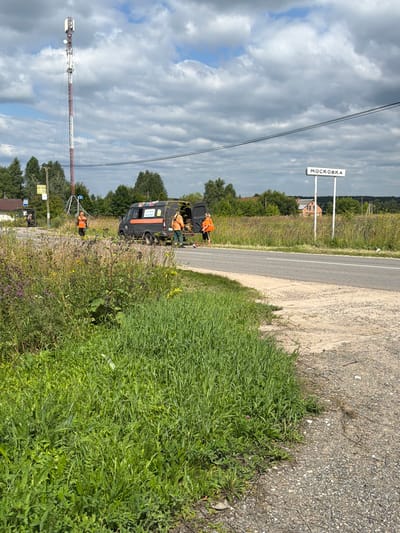 The image captures a roadside scene in Stankovo, Russia, on a cloudy day. In the foreground, a dense patch of green grass and wildflowers lines a gravel shoulder. A paved road runs through the mid-ground, with a dark grey van parked on its right side, its rear doors open. Three individuals wearing bright orange vests and dark pants are visible around the van. Two are standing near the open rear of the van, appearing to be working or interacting with the contents. Another individual in an orange vest stands further back, facing away from the camera. To the left of the van, a tall communication tower with several antennas is prominently featured. A white street sign with the Cyrillic text "МОСКОВКА" is mounted on a post to the right of the road. In the background, scattered trees and a few houses are visible amidst a field of tall grass. The overall impression is one of a roadside service or maintenance activity taking place in a rural or semi-rural setting.