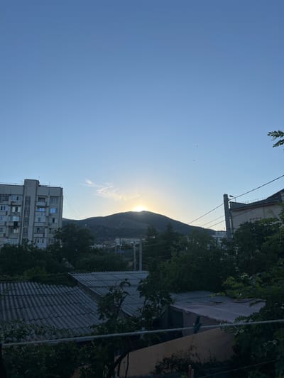 This is a vertically oriented, outdoor, daytime photograph captured in Sudak, Ukraine. The image depicts a scene with a clear blue sky dominating the left portion, sparsely populated with a few small birds. On the right, a mountain looms behind the horizon, partially obscuring the setting sun, which casts a warm glow across the sky.
In the foreground, an apartment building with multiple windows is visible, suggesting an urban or semi-urban setting. Overgrown trees and foliage frame the right side of the image, some of which appear to be leaning over buildings. A gray, tiled roof of a structure is prominent in the lower right corner, partially obscured by dark green leaves. Electrical wires crisscross the sky in the mid-ground, connecting to a utility pole. The overall impression is one of a quiet, everyday scene in a Mediterranean-like coastal town, possibly during the late afternoon or early evening as indicated by the low angle of the sun. There is no visible text in the image.