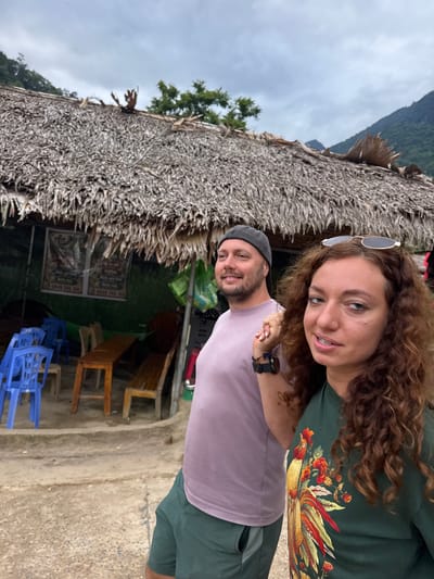 A man and a woman are standing in front of a thatched-roof structure in Cổ Lũng Commune, Vietnam. The man, wearing a gray beanie and a lavender t-shirt, is on the left side of the frame, smiling. The woman, with curly brown hair and sunglasses perched on her head, is on the right, looking at the camera. She is wearing a green t-shirt with a colorful rooster embroidered on it. Behind them, there are blue plastic chairs and wooden tables, suggesting an outdoor seating area, possibly a cafe or restaurant. The thatched roof is made of dried leaves and branches. In the background, lush green mountains are visible under a cloudy sky. The ground is a dirt path.