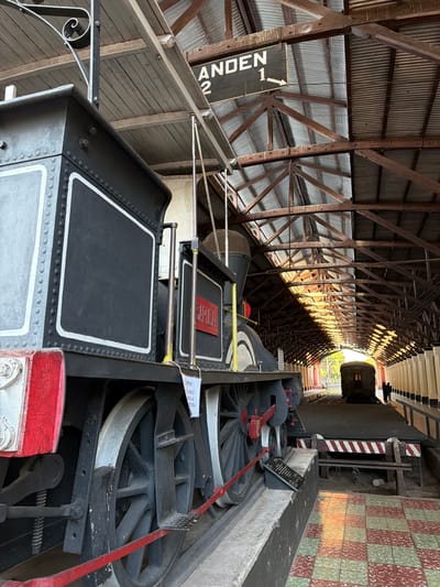 The image shows a steam locomotive and a covered train station. The locomotive is black with white trim and red accents. It has large wheels and is on display. A sign above the train reads "ANDEN 2 1" with an arrow pointing to the right. The train station has a corrugated metal roof supported by wooden beams. The floor is tiled in a red and green checkerboard pattern. In the background, another train car is visible. Two people are standing on the platform in the distance. The scene is likely the Asunción Railway Museum or a similar historical site in Asunción, Paraguay, given the context of preserved locomotives and vintage stations. The sunlight streams into the station, creating patterns of light and shadow on the floor and the structure.