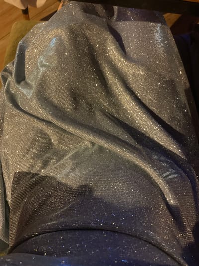 The image shows a close-up of a shimmering, metallic fabric, likely a dress or a piece of clothing, draped over a surface. The fabric is a silvery-blue color with many small, reflective sparkles. The way it is folded creates deep shadows and highlights, emphasizing its texture and sheen. The background is out of focus but suggests an indoor setting, possibly a domestic environment. There is no identifiable information about the city of Hadera, Israel, within the image.