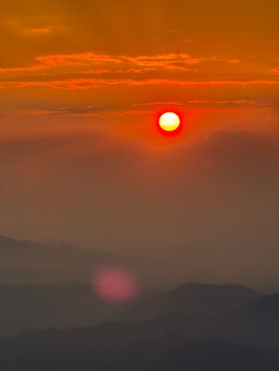 The original image presents a striking, vertically oriented view of a sunset or sunrise over a hazy landscape, characterized by warm, intense colors transitioning to cooler, darker tones. On the left side of the frame, a bright, circular yellow sun, surrounded by a red glow, is the focal point, casting its light through a thick atmosphere. Adjacent to the sun, the sky is filled with prominent vertical streaks and patches of orange and red, indicative of clouds or dense atmospheric haze illuminated by the low sun. Towards the right, the scene transitions dramatically into a darker, more muted area of deep grey and brown, which appears to be a silhouette of distant mountains or hills shrouded in heavy haze or shadow. A blurred, circular red-pink light anomaly, possibly a lens flare, is visible in the lower right portion of the image. The overall impression is a powerful, dramatic, and potentially smoky or polluted sky at dawn or dusk, possibly within the Baiyun District of China. There is no visible text in the image.