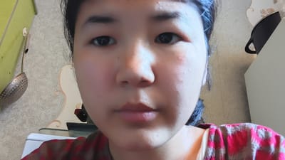 The image is a close-up selfie of a young woman. She has dark hair pulled back from her face, with a few strands falling forward. Her eyes are dark and her eyebrows are dark and well-defined. Her nose is small and her lips are full and pink. She is wearing a red and brown plaid shirt. The background appears to be a domestic setting, with a patterned wall and some kitchen utensils visible. There is no identifiable information to place this image in the city of Uchkun, Kyrgyzstan.