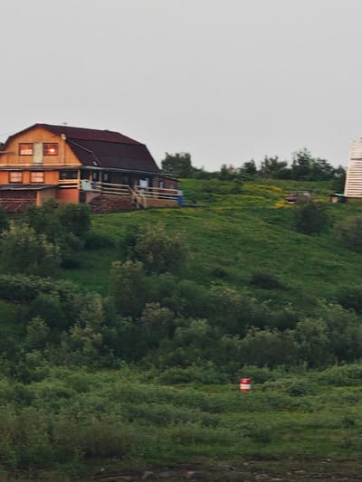 The image depicts a rural scene in Potapovo, Russia, under an overcast sky, suggesting it's either dawn or dusk. The primary subject is a multi-story wooden house situated on a grassy hillside, with a dark-colored roof. The house has several windows, some of which are illuminated from within, casting a warm glow. A wooden deck with railings surrounds a portion of the house.
The hillside is covered in lush green grass, dotted with patches of yellow wildflowers and interspersed with various trees and bushes. A red and white striped barrel stands on the lower part of the hill. In the distance, atop the hill, a red vehicle is parked near a small structure, and further to the right, a tall, white, slatted structure, possibly a tower or a windmill, is partially visible. Another small structure is located to the right of the red vehicle.
The overall mood is tranquil and somewhat isolated, characteristic of a remote countryside location. The soft, diffused light indicates a lack of direct sunlight, reinforcing the impression of early morning or late evening. There is no visible text in the image.