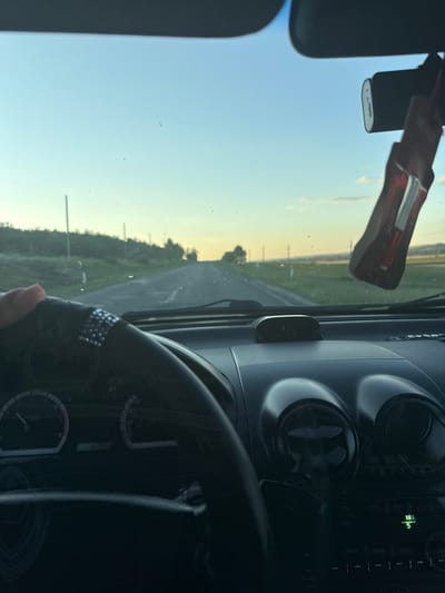 The image shows a first-person perspective from inside a vehicle, likely in Iordanovca, Moldova, given the context. The vehicle appears to be moving along a paved road bordered by grassy fields and scattered trees. The sky is a gradient of pale blue to a soft yellow near the horizon, suggesting either dawn or dusk.
In the foreground, a person's left hand is visible gripping the steering wheel, which is covered in black material and decorated with a band of rhinestones. The dashboard is visible, featuring the steering wheel, an instrument cluster with a tachometer on the left and a speedometer on the right. A small digital display showing "8:13" is mounted on the dashboard above the radio.
A dashcam is mounted on the windshield, directly above the rear-view mirror. Hanging from the rear-view mirror is a car air freshener, likely red in color with some white lettering. The windshield shows some dust or smudges. Electrical wires are visible near the top edge of the windshield, possibly related to the dashcam. The overall impression is a serene drive through a rural landscape during a transitional time of day.