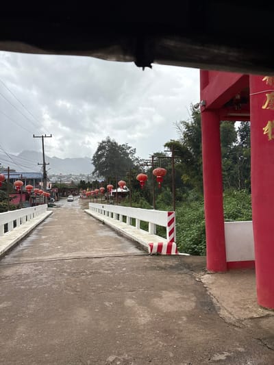 An image captures a wet, concrete road and bridge in Ban Mai Saha Sampan, Thailand, on an overcast day. The perspective is from beneath a dark, indistinct overhead structure, possibly the interior roofline of a vehicle.
In the immediate foreground, a rough concrete road surface is visible, leading to a concrete bridge. On the right, a red and white striped barrier marks the approach to the bridge, adjacent to a large, red archway or gate structure with gold-colored script on its visible column. Lush green vegetation grows densely on the right side beyond this archway.
The bridge features white concrete balustrades along both sides. The road continues slightly uphill beyond the bridge, narrowing into the distance. Numerous red, spherical Chinese lanterns hang from dark poles that line both sides of the road and bridge, extending into the background.
A white sedan is visible on the road in the mid-distance, positioned centrally. Further in the background, a cluster of residential or commercial buildings with varied roofs lines the road. Utility poles with overhead wires are present. Distant mountains, partially obscured by atmospheric haze or clouds, form the horizon under a heavily clouded sky. No individuals are visible in the scene.