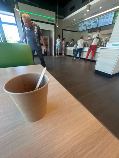In Polsko Padarevo, Bulgaria, a man wearing a dark t-shirt with graphics and black pants is standing near a green booth and looking down at a tablet. The man has short, graying hair. In the background, several people are lined up near a counter with a menu displayed on screens above. A brown paper cup with a wooden stirrer stands on a wooden table in the foreground. The environment appears to be an indoor fast-food restaurant or a similar establishment, with tiled flooring and a bright, modern interior.