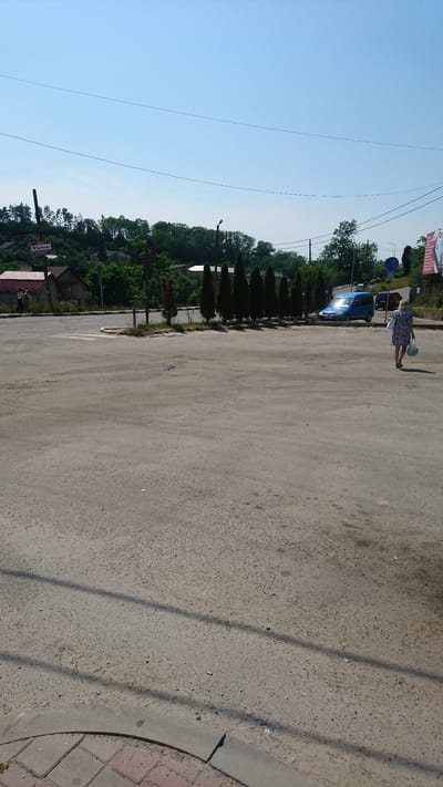 A full outdoor, eye-level shot captured on a sunny day shows an open concrete area with a few cypress trees planted in a row. A blue minivan is parked to the right of the trees, and a woman in a floral dress walks away from the camera, carrying a bag. In the background, a line of trees and houses is visible, with a billboard on the right side of the frame. The scene appears to be a public space, possibly a parking lot or a town square, in Buchach, Ukraine.